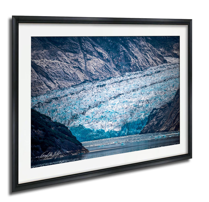 A framed photograph depicting a large, blue-green glacier with ice formations and a body of water in the foreground.