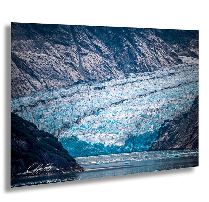 A large, majestic glacier with a blue-green hue dominates the scene, partially submerging the surrounding landscape. The glacier extends across the entire image, creating a striking contrast against the rocky terrain. The water below the glacier appears calm and serene, reflecting the natural beauty of the scene. The image is a captivating representation of the power and grandeur of nature.