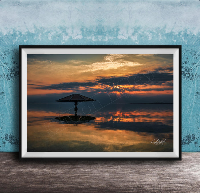 A framed photograph depicting a serene sunset over a calm body of water, with a lone umbrella standing in the foreground.