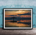 A framed photograph depicting a serene sunset over a calm body of water, with a lone umbrella standing in the foreground.