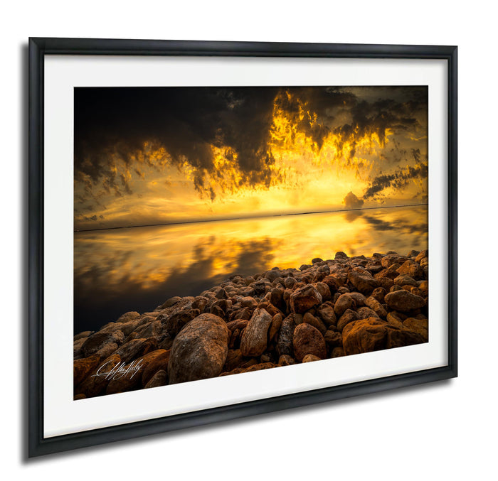 The image is a framed photograph depicting a rocky shoreline with a dramatic sunset in the background.