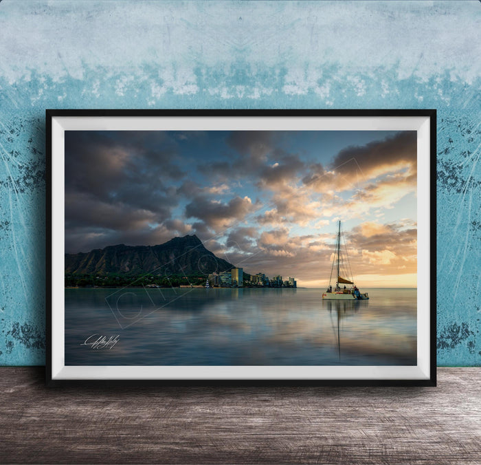 A framed photograph depicting a serene scene of a sailboat on a calm body of water, with a mountainous landscape and a colorful sunset in the background.