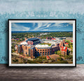 The image is a framed aerial view of a large sports stadium, showcasing its distinctive architecture and surrounding landscape.
