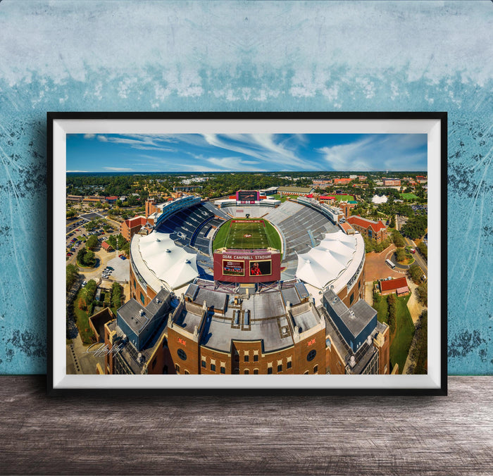 The image is an aerial view of a large sports stadium with a football field, surrounded by buildings and a blue sky.