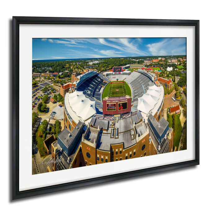 The image is an aerial view of a large sports stadium with a football field, surrounded by buildings and greenery.
