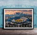 An aerial view of a baseball stadium with the city skyline in the background, framed in a black frame and displayed on a wooden surface.