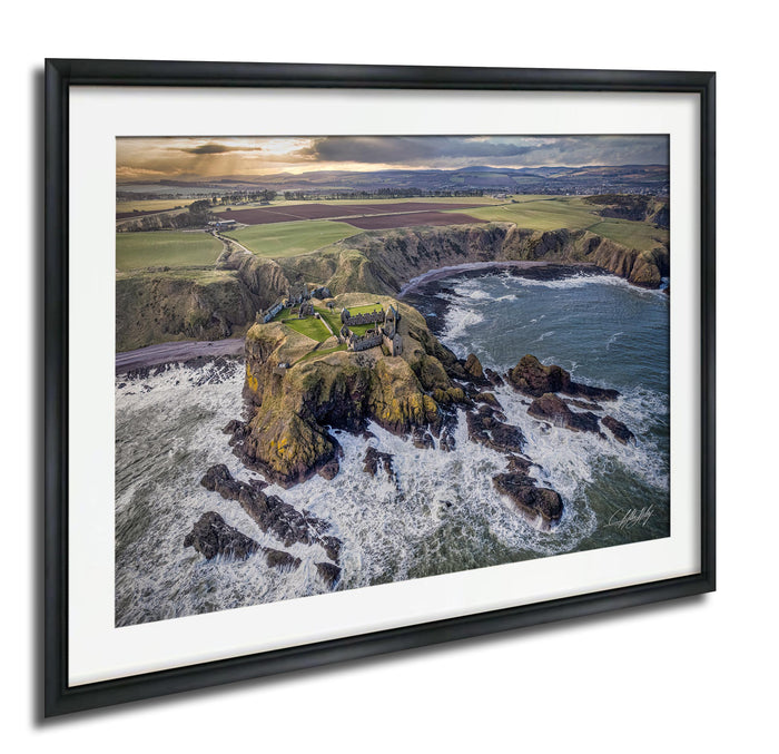The image is a framed aerial view of a coastal landscape, featuring a rocky outcrop, a small village or settlement, and the ocean with crashing waves.