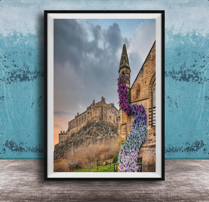 A framed photograph of a castle with a tower, set against a cloudy sky, with a colorful flower arrangement hanging from the tower.