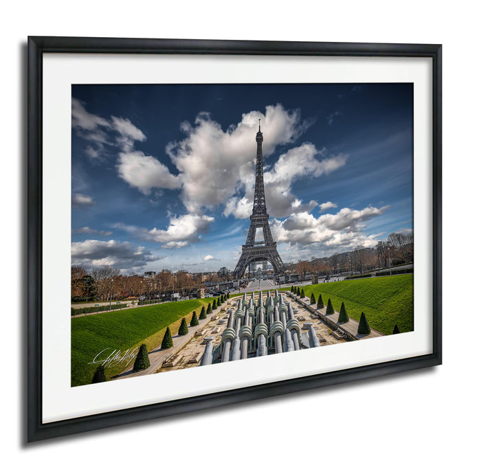 the Eiffel Tower in Paris, France, with a cloudy blue sky and lush green gardens in the foreground.