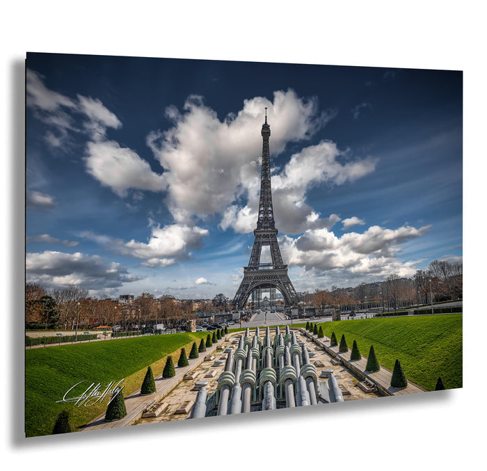 the Eiffel Tower in Paris, France, with a cloudy blue sky in the background. The tower is surrounded by a well-maintained garden with neatly trimmed hedges and trees. The overall scene is visually appealing and captures the iconic landmark.