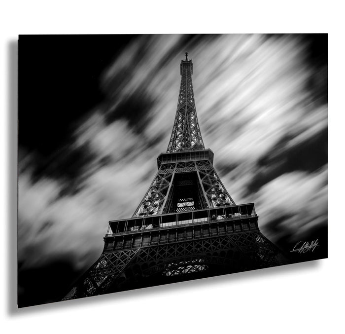 The image is a black and white photograph of the Eiffel Tower, a famous landmark in Paris, France. The tower is captured from a low angle, emphasizing its height and grandeur. The sky in the background is filled with clouds, creating a dramatic and moody atmosphere. The image is a collage of multiple images, and the artist's signature is visible in the bottom right corner.