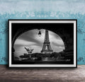 A black and white photograph of the Eiffel Tower in Paris, France, framed and displayed on a wooden surface against a blue textured wall.