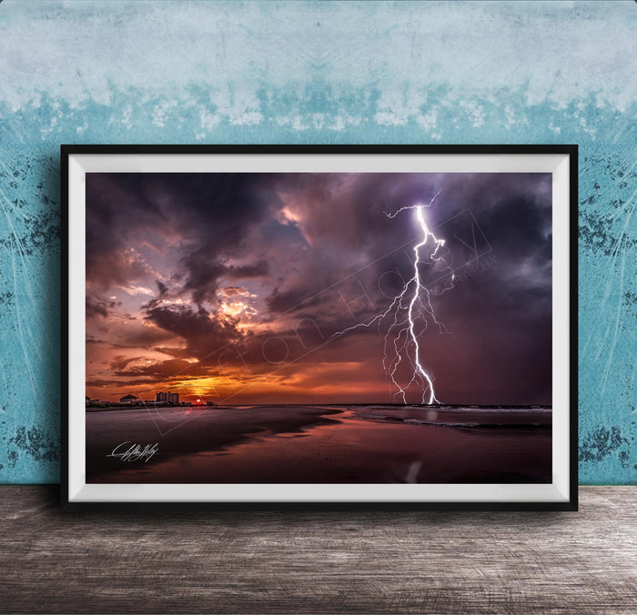 A framed photograph depicting a dramatic lightning storm over a beach at sunset, with the sky filled with vibrant colors and the lightning bolt striking the horizon.