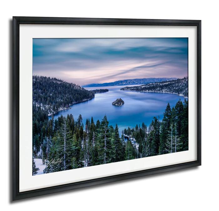 A framed photograph depicting a serene landscape with a lake surrounded by a forested area and mountains in the distance.