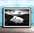 A black and white photograph of a serene lake surrounded by a forested landscape, with a cloudy sky above.