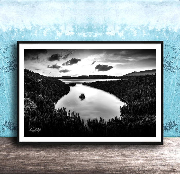 A black and white photograph of a serene lake surrounded by a forested landscape, with a cloudy sky above.