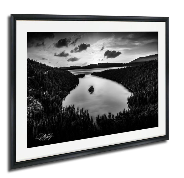A black and white landscape photograph depicting a serene lake surrounded by a forested area, with mountains visible in the distance under a cloudy sky.
