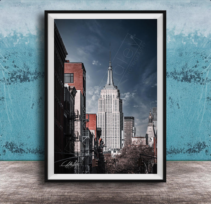 A framed photograph of the iconic Empire State Building in New York City, with a blue sky and clouds in the background.