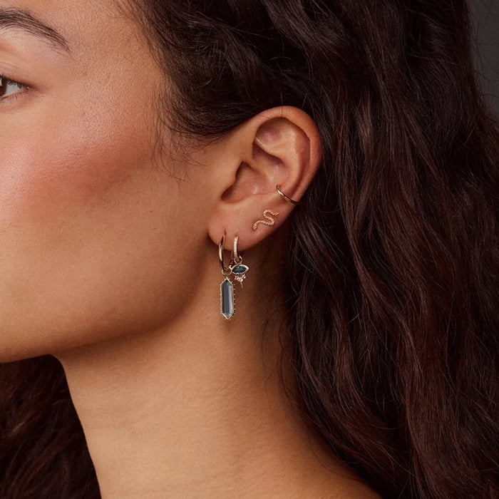 Close-up of a model's ear wearing a gold huggie hoop earring with a dangling london blue topaz stone and a small gold snake ear cuff.