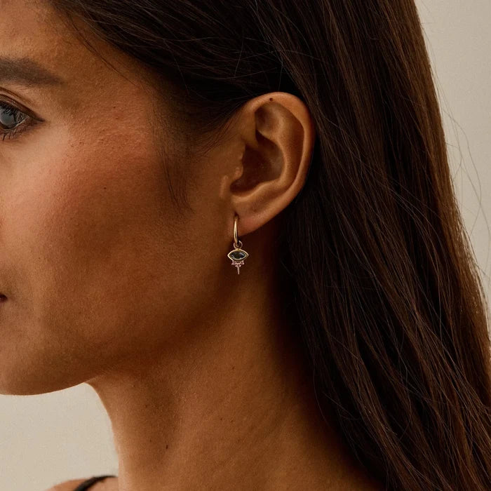 A close-up side profile of a woman wearing a delicate solid gold huggie hoop earring with a small evil eye earring charm featuring london blue topaz stone