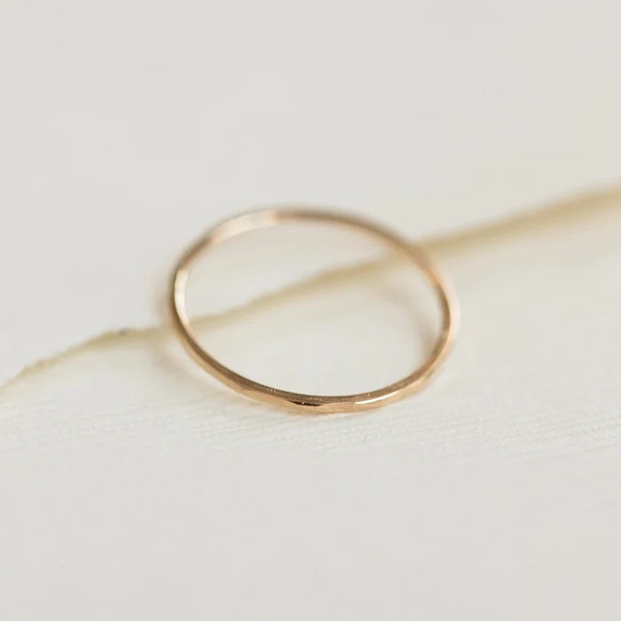 A delicate **Faceted Felicity Stacking Ring - 10k Yellow Gold** from *bluboho* sits on a light-colored, textured surface. The ring features a sleek, smooth design with soft lighting highlighting its minimalistic elegance.