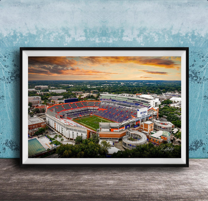 The image is a framed photograph of a large sports stadium, likely a football stadium, with a colorful sunset sky in the background.