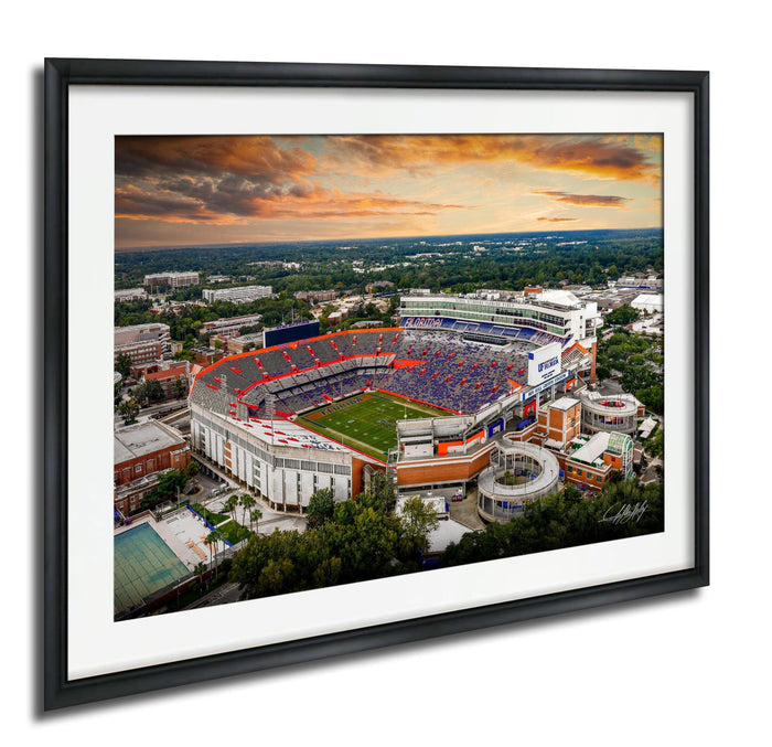 An aerial view of a large sports stadium with a crowd of spectators, surrounded by a cityscape and a dramatic sky with clouds and colors.