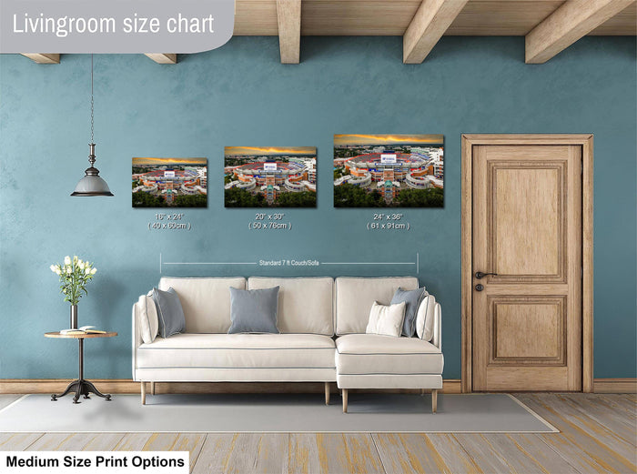 The image is a livingroom size chart with three framed prints of a stadium or sports arena displayed on the wall. The prints vary in size, ranging from 10'x12' to 24'x36'', and are positioned above a white couch. The livingroom has a wooden door and a small table with a vase of flowers, creating a cozy and inviting atmosphere.