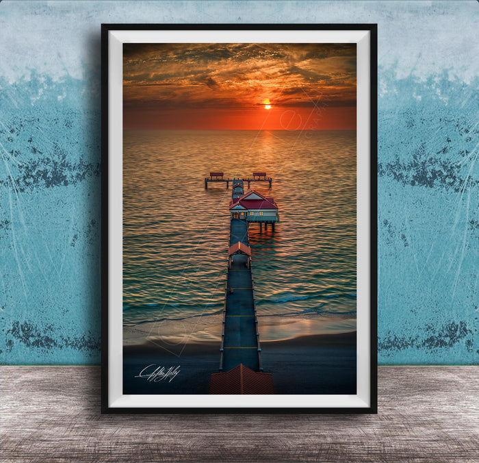 A framed photograph depicting a serene sunset over a body of water, with a pier extending into the water and a small building on the pier.