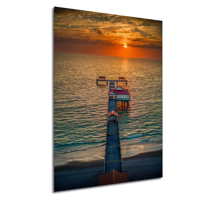 a long pier extending into a calm body of water, with a red building on the pier and a vibrant sunset in the background.