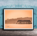A framed photograph of a wooden pier extending into a body of water, with a warm orange and yellow sunset sky in the background.