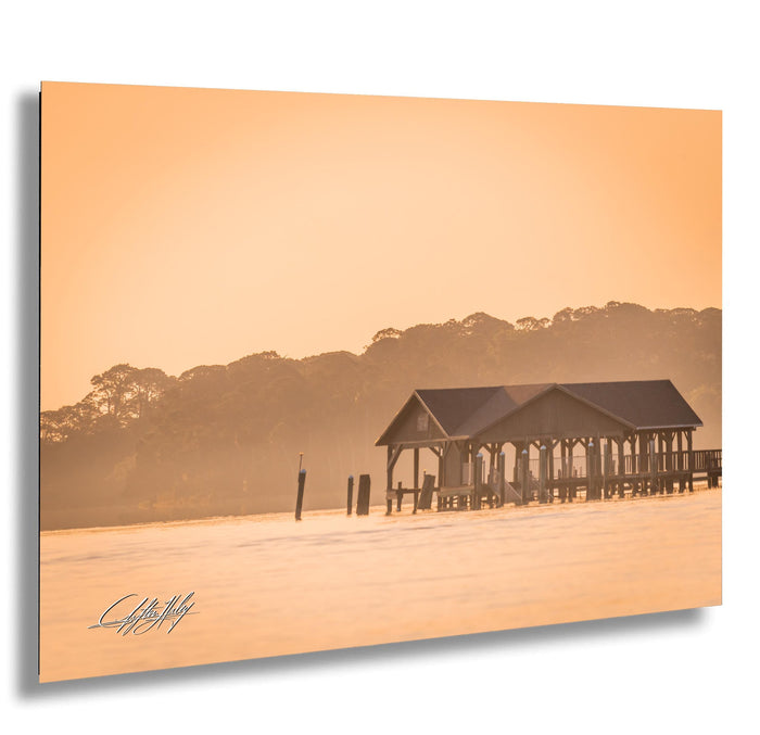 A wooden pier extends into a body of water, with a small building on the pier and a line of trees in the background. The sky is hazy, creating a warm, golden glow over the scene.