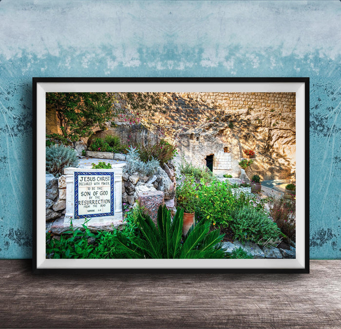 A framed photograph depicting a stone wall with a sign that reads "JESUS CHRIST" and "SON OF GOD". The wall is surrounded by lush greenery, including plants and trees.