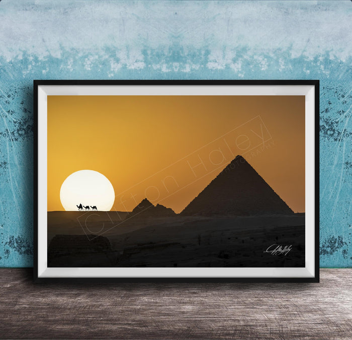 A framed photograph depicting the silhouette of the Great Pyramids of Giza against a vibrant sunset sky, with camels in the foreground.