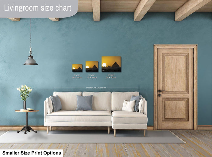 The image is a living room with a white sofa, a wooden door, and a hanging light fixture. There are three framed pictures of sunsets displayed on the wall above the sofa.