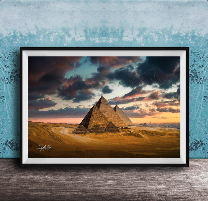 A framed photograph of the iconic pyramids of Giza against a dramatic sky at sunset, with the sun setting behind the sand dunes.