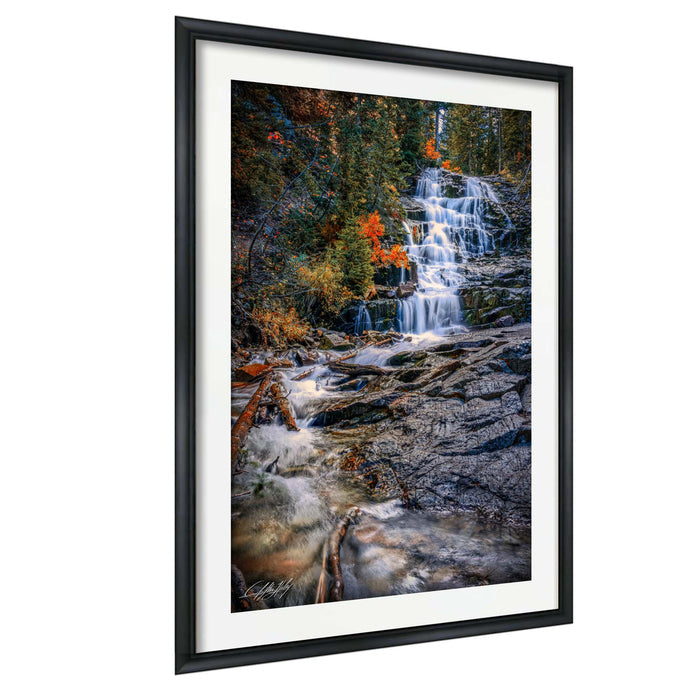 A framed photograph depicting a waterfall cascading down a rocky cliff surrounded by trees with autumn foliage.