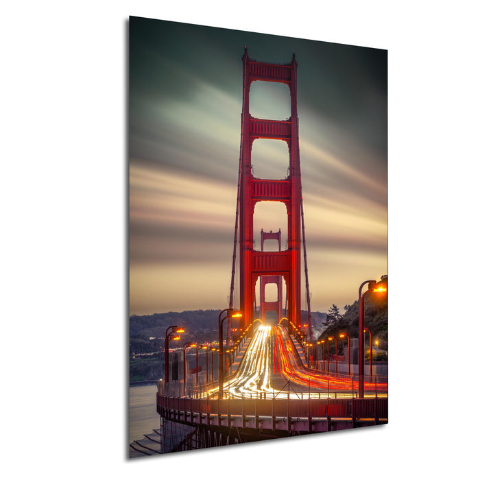 a large suspension bridge with a distinctive red color, spanning across a body of water at sunset. The bridge is illuminated by warm, glowing lights, creating a picturesque scene. The sky above is filled with clouds, adding to the dramatic atmosphere of the image. The bridge's lights are reflected on the water below, creating a mesmerizing visual effect.