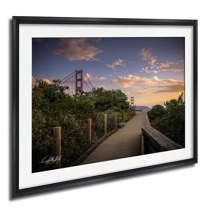 a scenic view of the Golden Gate Bridge at sunset, with the bridge's distinctive red towers silhouetted against a colorful sky. The bridge is surrounded by lush greenery, and a wooden walkway leads up to it. The warm hues of the sunset create a serene and picturesque atmosphere, highlighting the beauty of the landmark.