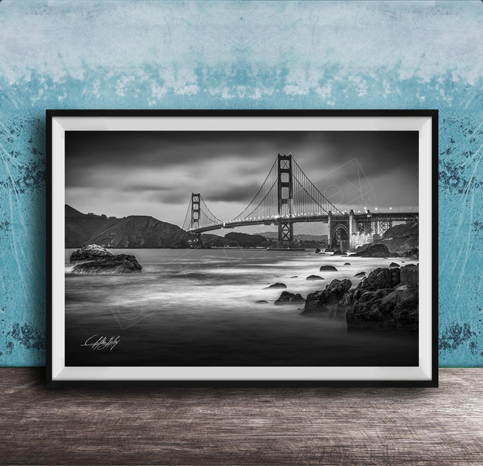 A black and white photograph of the Golden Gate Bridge in San Francisco, California, with the bridge's towers and suspension cables prominently featured against a cloudy sky.