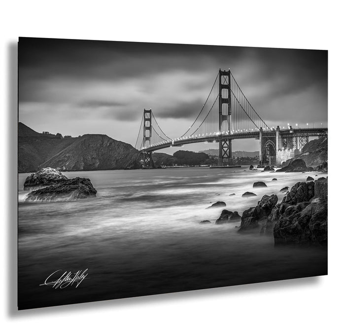 A black and white photograph of the Golden Gate Bridge in San Francisco, California, with the ocean and rocky coastline in the foreground.