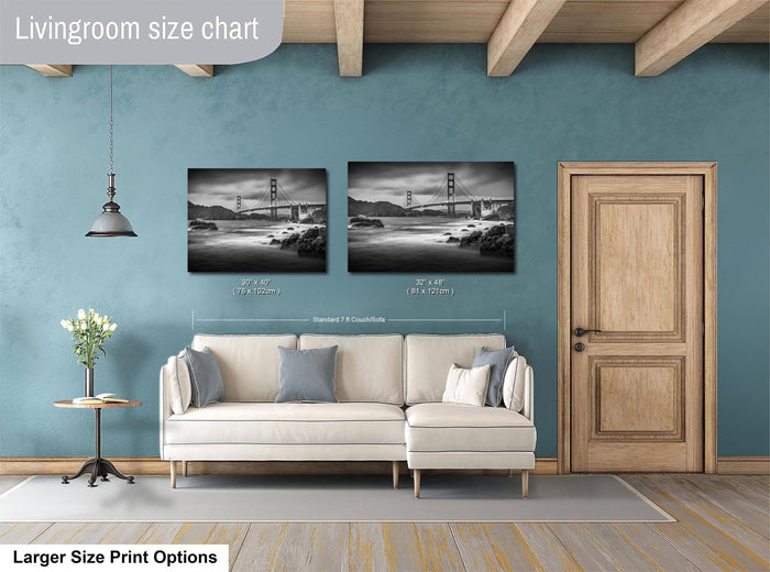 a living room with a white sofa and a wooden door. Above the sofa, there are two framed black and white photographs of the Golden Gate Bridge. The wall behind the sofa has a blue color, and there is a lamp hanging from the ceiling. The living room appears to be well-lit and spacious.