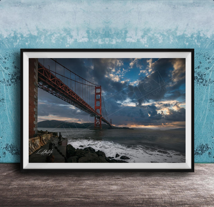 A framed photograph of the Golden Gate Bridge in San Francisco, California, with dramatic clouds and a sunset sky in the background.