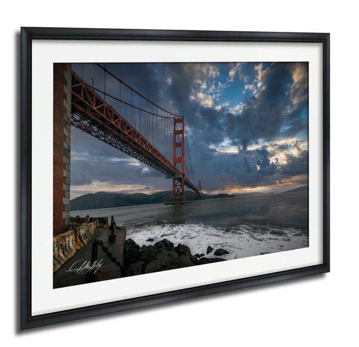 a large suspension bridge spanning a body of water, with dramatic clouds and a sunset sky in the background.