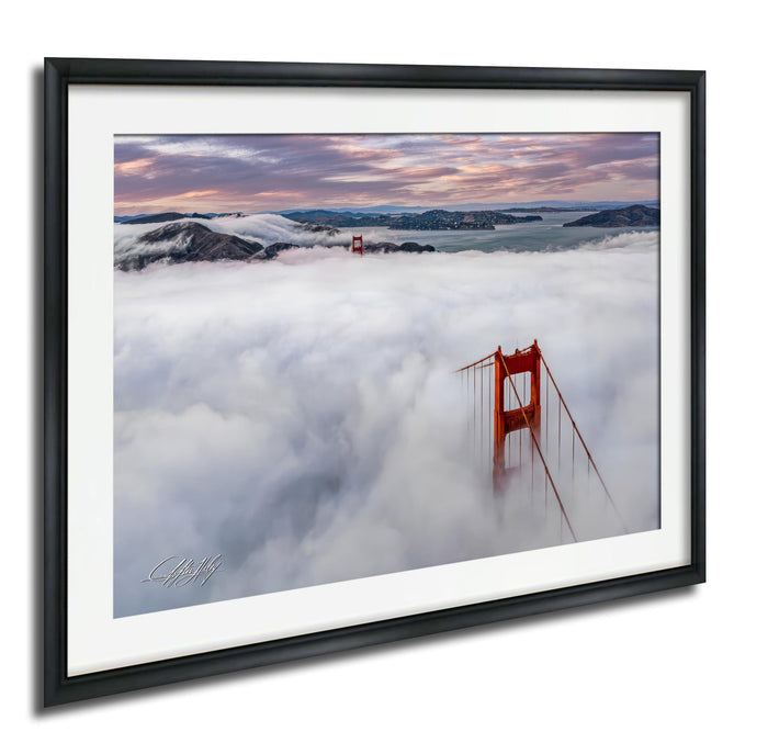 a suspension bridge, the Golden Gate Bridge, standing tall amidst a sea of clouds. The bridge's distinctive red color contrasts with the surrounding white fog, creating a striking visual effect. The photograph is framed and displayed on a wall, capturing the beauty of the iconic landmark.