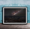 The image is a framed photograph of a night sky with a galaxy, placed on a wooden surface against a blue wall.