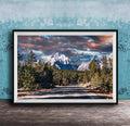 A framed photograph depicting a scenic mountain landscape with a winding road surrounded by pine trees and snow-capped peaks under a dramatic cloudy sky.