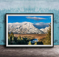 A framed photograph of a mountainous landscape with snow-capped peaks, a river, and a forested area in the foreground.