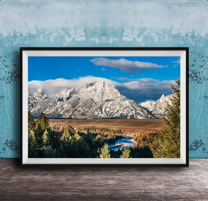 A framed photograph of a mountainous landscape with snow-capped peaks, a river, and a forested area in the foreground.