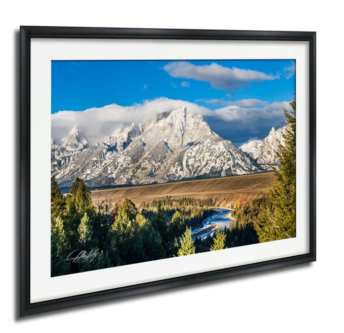 A framed photograph depicting a scenic mountain landscape with snow-capped peaks, a winding river, and a mix of evergreen and deciduous trees.
